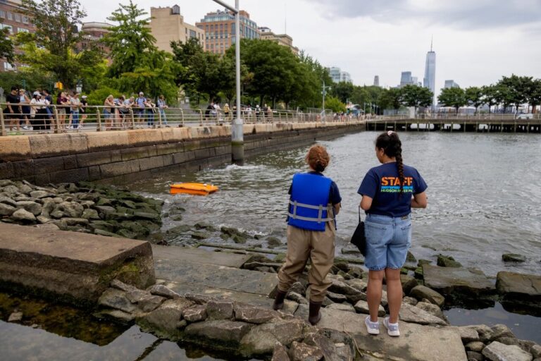 An orange WasteShark in the Hudson River, two people controlling its movements, spectators watching on in front of New York City