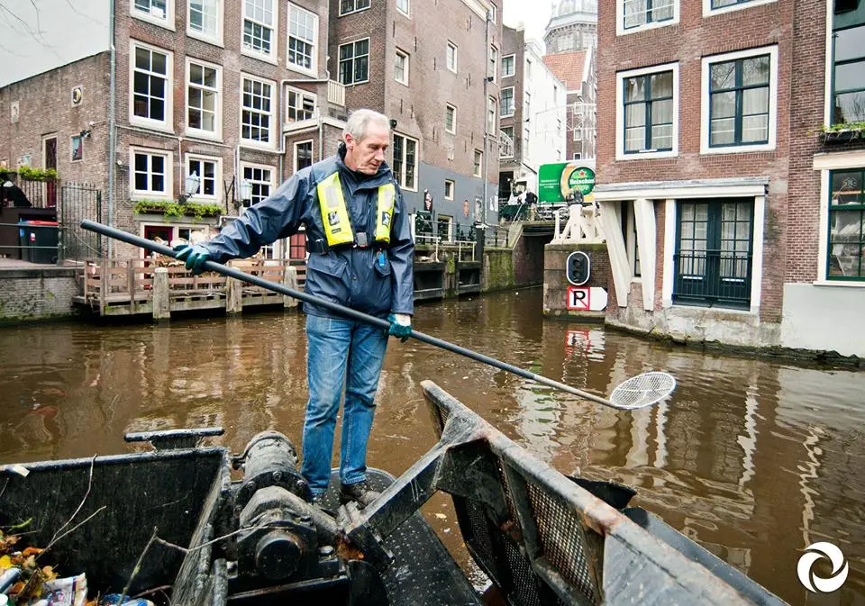 A person cleaning the canals n Amsterdam by hand on a boat. Picture is used to show the cost for manual labor versus the use of a RanMarine WasteShark Plus.