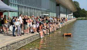 People sitting and standing next to a waterway inside a city watching a WasteShark Plus coming by. 